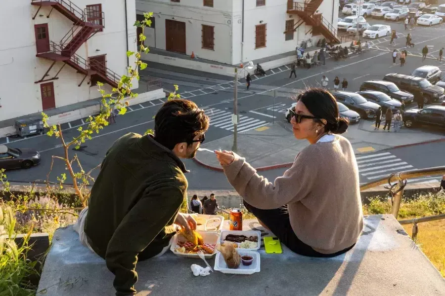 Um casal janta ao ar livre no Fort Mason Center em São Francisco. A mulher alimenta seu companheiro com um gole de comida.